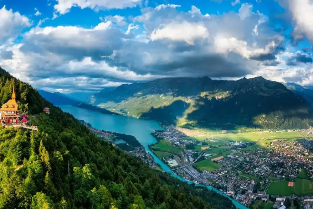 Vista panorámica de Interlaken, Suiza, con el lago Brienz y montañas rodeadas de frondosos bosques bajo un cielo parcialmente nublado.