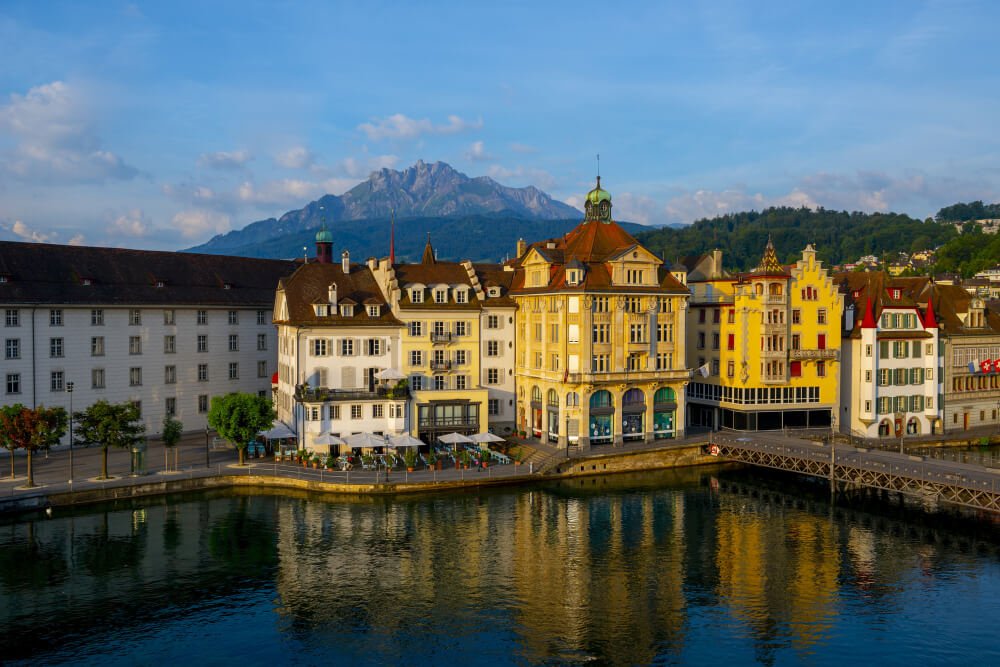 Vista panorámica de edificios históricos junto al río Reuss en Lucerna, Suiza, con el monte Pilatus al fondo en un día soleado.