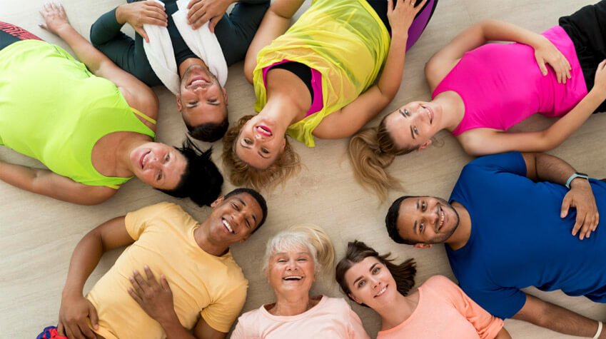 Grupo de personas de diversas edades y géneros acostadas en círculo sobre el suelo, todas sonrientes y vistiendo ropa deportiva de colores brillantes. _Crédito a Freepik