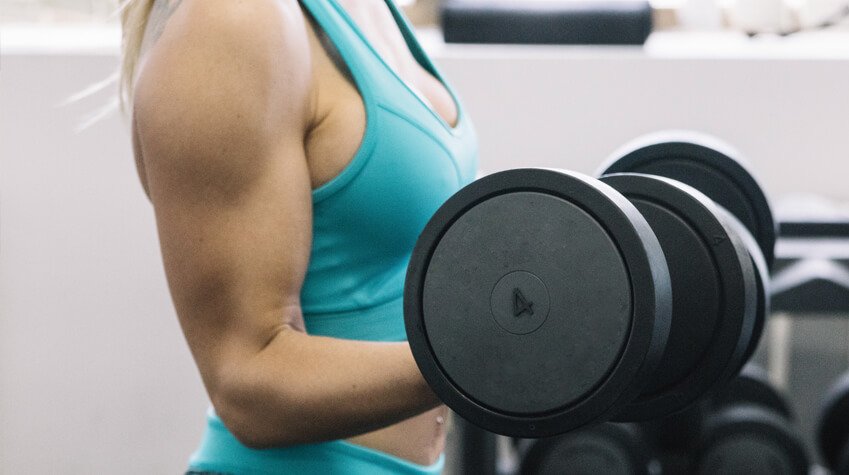 Mujer con top deportivo turquesa levantando una mancuerna negra en el gimnasio, mostrando fuerza y definición muscular en su brazo. Ganar Músculo _ crédito freepik