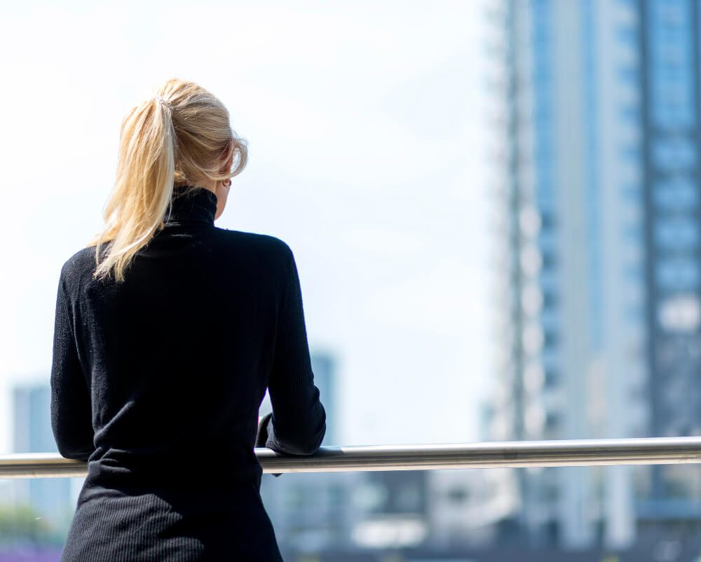 Mujer rubia con coleta, de espaldas, observando la ciudad desde un balcón en un entorno urbano. Imagen cortesía de Freepik.