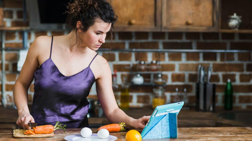Mujer en la cocina usando una tablet mientras corta zanahorias en una tabla de madera. En la encimera también hay huevos y un limón, sugiriendo que está siguiendo una receta o plan de cocina saludable para Ganar Músculo. _Crédito freepik